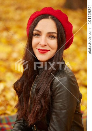 Beautiful woman in autumn park, enjoing warm sunny weather. Close up image of happy dark haired young wearing black leather jacket and red beret, looking with charming smile directly at camera. Beautiful woman in autumn park, enjoing warm sunny weather. Close up image of happy dark haired young wearing black leather jacket and red beret, looking with charming smile directly at camera. 101139294