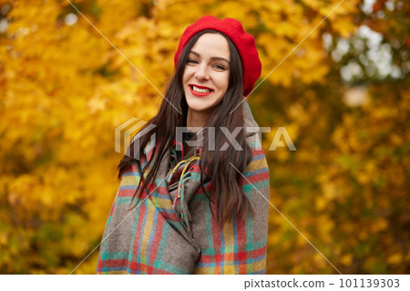 Young beautiful woman smiling female, wearing fashionable red beret and wrapped checkered blanket posing isoalted over nature background. Outdoor portrait of attarctive happy girl in autumn. 101139303