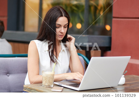 Portrait of concentrated brunette female works on business project while uses modern laptop computer, focused into screen, drinks beverage, sits against coffee shop interior, messages with friends 101139329