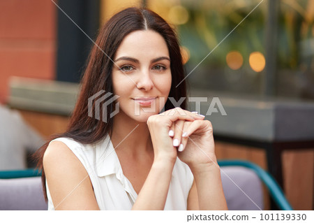 Close up shot of pretty brunette young female with dark hair, healthy skin, holds hands under chin, looks mysteriously at camera, has pleasant talk with friend, poses against blurred background 101139330