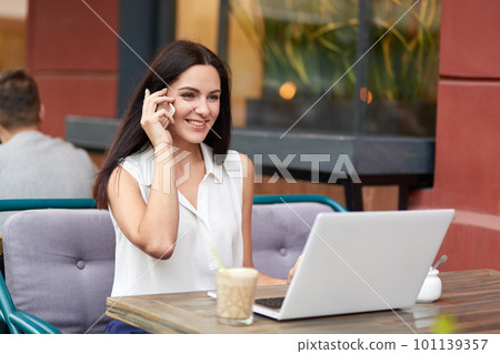 Positive businesswoman in formal clothes, has telephone conversaton, speaks with colleague, uses laptop computer for making business report, drinks milk cocktail, poses in outdoor restaurant Positive businesswoman in formal clothes, has telephone conversaton, speaks with colleague, uses laptop computer for making business report, drinks milk cocktail, poses in outdoor restaurant 101139357