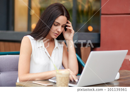 Horizontal shot of pretty busy female freelancer sits in front of opened laptop, drinks coffee, works on new project, surrounded with modern technologies, wears white blouse. sits at outdoor cafe Horizontal shot of pretty busy female freelancer sits in front of opened laptop, drinks coffee, works on new project, surrounded with modern technologies, wears white blouse. sits at outdoor cafe 101139361