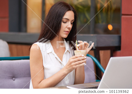 Beautiful brunette female student works on scientific project via laptop computer, searches online, dressed in elegant white blouse, hold glass of cocktail, spends free time in outdoor cafeteria. Beautiful brunette female student works on scientific project via laptop computer, searches online, dressed in elegant white blouse, hold glass of cocktail, spends free time in outdoor cafeteria. 101139362
