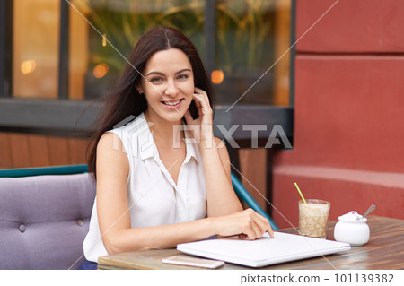Positive dark haired woman in white outfit, surrounded with modern gadgets, sits at coffee shop, smiles gently at camera, being in good mood, waits for order, going to gave lunch after work. Positive dark haired woman in white outfit, surrounded with modern gadgets, sits at coffee shop, smiles gently at camera, being in good mood, waits for order, going to gave lunch after work. 101139382