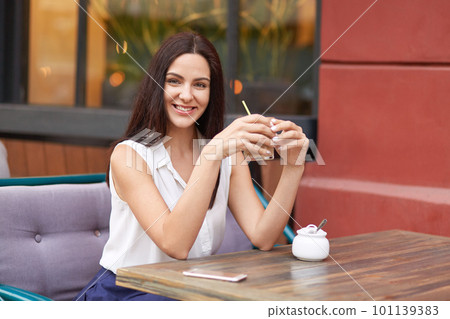 Pleased brunette lady holds fresh cocktail, sits at wooden table in cozy outdoor cafeteria, uses modern mobile phone, spends spare time with friends, has day off. People, leisure, lifestyle concept Pleased brunette lady holds fresh cocktail, sits at wooden table in cozy outdoor cafeteria, uses modern mobile phone, spends spare time with friends, has day off. People, leisure, lifestyle concept 101139383