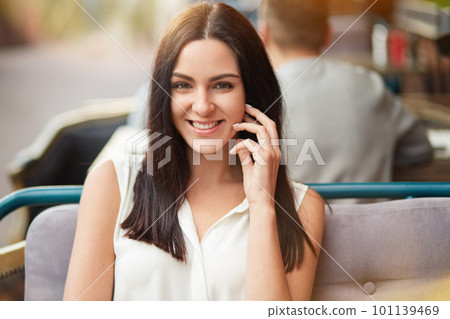 People, beauty and happiness concept. Attractive brunette female in white blouse, has delighted expression, healthy skin, sits against blurred outdoor cafe background, smiles broadly at camera People, beauty and happiness concept. Attractive brunette female in white blouse, has delighted expression, healthy skin, sits against blurred outdoor cafe background, smiles broadly at camera 101139469