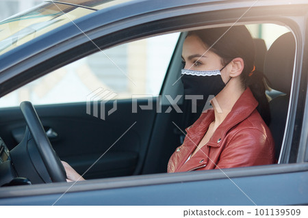 Portrait of attentive focused cute young brunette wearing protective mask, sitting in car, being in traffic jam, opening window, being alone, caring about her health. Coronavirus outbreak concept. 101139509