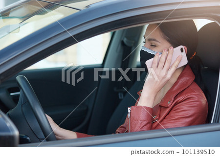Horizontal picture of positive smiling young woman in mask talking over phone, holding smartphone in hands, driving car, being alone in vehicle, unsafety measures. People and quarantine concept. Horizontal picture of positive smiling young woman in mask talking over phone, holding smartphone in hands, driving car, being alone in vehicle, unsafety measures. People and quarantine concept. 101139510