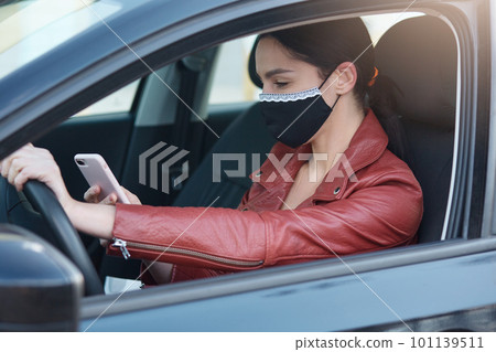 image of girl with dark hair and ponytail, dresses leather jacket and protective mask,holding phone in hands, checking social network or call number while sitting at driver's seat in automobile. 101139511