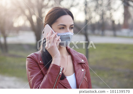 Portrait of thoughtful serious brunette holding smartphone near ear, wearing medical mask, talking over phone, being alone outdoors, using device, breaking quarantine rules. Covid19 concept. 101139570
