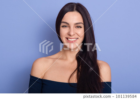 Portrait of young beautiful cute cheerful girl, stands smiling and looking at camera, posing isolated over blue background, attractive lady with bare shoulders, expresses happyness, being in good mood Portrait of young beautiful cute cheerful girl, stands smiling and looking at camera, posing isolated over blue background, attractive lady with bare shoulders, expresses happyness, being in good mood 101139584