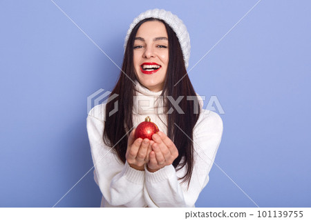 Image of energetic joyful brunette smiling sincerely, having pleasant facial expression, being in high spirits, enjoying new year holidays. holding red christmas toy in both hands. Rest time concept. 101139755