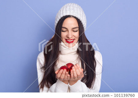 Girl puts her hands together and Christmas tree bauble lies on them, brunette female wearing warm white clothing looking at her palms with christmas ball, adorable woman expressing happyness. 101139760