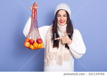 Photo of cheerful good looking adorable young woman holding handbag with fruit, directing with forefinger at it, standing isolated over lilac background, laughing sincerely. Winter time concept. 101139768