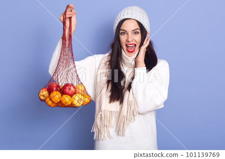 Excited young woman with fresh ripe mandarin, orange fruit isolated over blue background in studio, female wearing white winter cap and sweater, posing with opened mouth, keeps hand on cheek. Excited young woman with fresh ripe mandarin, orange fruit isolated over blue background in studio, female wearing white winter cap and sweater, posing with opened mouth, keeps hand on cheek. 101139769