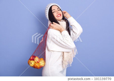 Horizontal studio shot of funny positive brunette wearing white sweater, hat and scarf, holding handbag with apples and mandarines, looking directly at camera, enjoying winter holidays. New Year 101139770