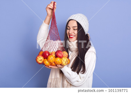 Portrait of cheerful good looking brunette looking at mandarines and apples, holding net handbag in hands, being in good mood, fond of fruit, spending time alone, having red lips. New Year atmosphere 101139771
