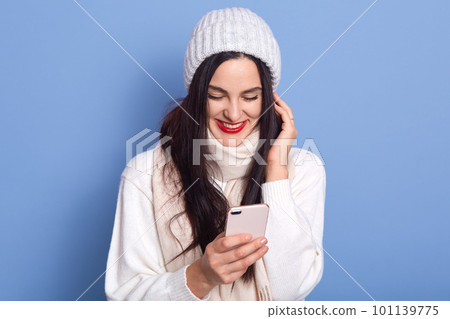 Portrait of nice attractive satisfied cheerful girl holding in hands and using cell phone, lady stands isolated over bright blue background, female wearing white pullover, scarf and cap. Portrait of nice attractive satisfied cheerful girl holding in hands and using cell phone, lady stands isolated over bright blue background, female wearing white pullover, scarf and cap. 101139775