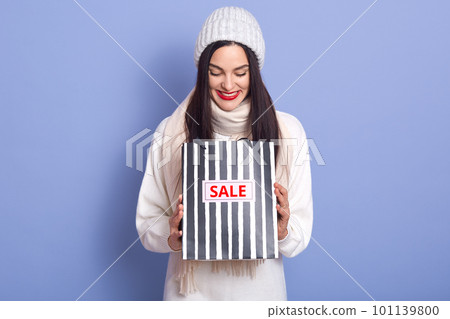 Curious woman with bright red lips and looking in her striped blackand whirte bag with inscription sale. Studio photo of emotional girl wearing white warm clothing isolated over blue background. Curious woman with bright red lips and looking in her striped blackand whirte bag with inscription sale. Studio photo of emotional girl wearing white warm clothing isolated over blue background. 101139800