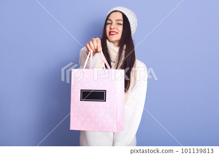 Portrait of beautiful pleasant female holding pink paper bag with purchase, looking directly at camera, having long black hair, spending time alone, enjoying winter holidays. Shopping concept. Portrait of beautiful pleasant female holding pink paper bag with purchase, looking directly at camera, having long black hair, spending time alone, enjoying winter holidays. Shopping concept. 101139813
