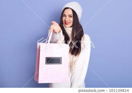 Indoor studio shot of cheerful beautiful female wearing white sweater, hat and scarf, holding paperbag with Christmas present, smiling sincerely, standing isolated over lilac background. Winter time. 101139814