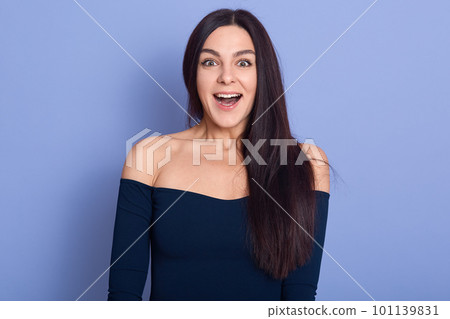 Close up studio photo of pretty charming cute female standing with open mouth, young smiling lady with long straight and bare shoulders, posing isolated over bright blue background. People concept. Close up studio photo of pretty charming cute female standing with open mouth, young smiling lady with long straight and bare shoulders, posing isolated over bright blue background. People concept. 101139831
