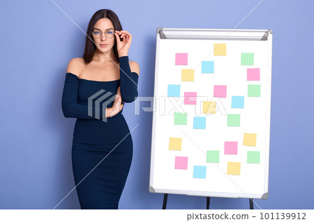 Front view of attractive woman wearing blue dress with bare shoulders, standing near white flipchart with multi colored stickers notes, holding hand on her spectacles. Copy space for advertisment. Front view of attractive woman wearing blue dress with bare shoulders, standing near white flipchart with multi colored stickers notes, holding hand on her spectacles. Copy space for advertisment. 101139912
