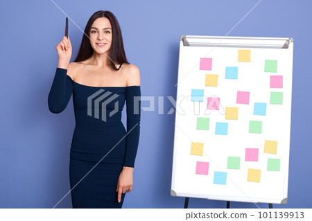 Young woman standing near sticky notes on flip chart, holding pen in raising hand, having great business idea, posing in dress isolated over blue background. Young woman standing near sticky notes on flip chart, holding pen in raising hand, having great business idea, posing in dress isolated over blue background. 101139913