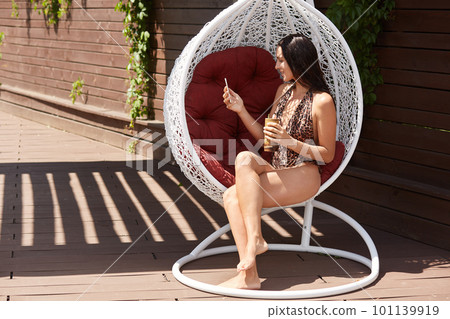 Brunette lady with wearing swimming suit with leopard print sitting on white stylish hanging chair and having video call or making selfie, enjoying drinking cocktail. 101139919