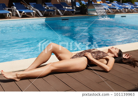 Image of attractive beautiful tanned woman relaxing in outdoor swimming pool in sun. Young female wearing bikini with leopard print laying near pool. Recreation, resting, summer time concept. 101139963