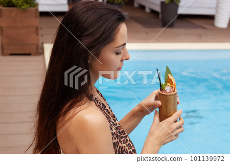 Priofile shot of pretty young slim stylish woman with long dark hair sitting near swimming pool with cocktail, wearing fashionable swimwear with leopard print. Concept of happy holiday and recreation. 101139972