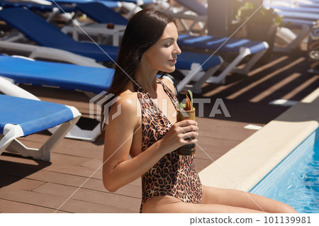 Woman with cocktail having fun near swimming pool, female with dark hair drink non alcoholic cocktail, wearing stylish swimming suit with leopard print, girl sunbathing and keeping eyes closed. Woman with cocktail having fun near swimming pool, female with dark hair drink non alcoholic cocktail, wearing stylish swimming suit with leopard print, girl sunbathing and keeping eyes closed. 101139981