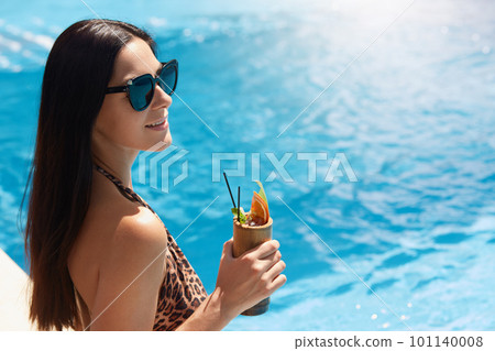 Happy young woman in dark sunglasses holding cocktail while standing next to swimming pool. Female in bikini with leopard print drinking fruit juice on summer day, spends summer days relaxing. Happy young woman in dark sunglasses holding cocktail while standing next to swimming pool. Female in bikini with leopard print drinking fruit juice on summer day, spends summer days relaxing. 101140008
