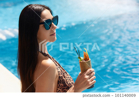 Tenned beautiful woman in leopard print swimming suit and black sunlasses sitting in swimming poolside edge with cocktail in hand. Fashionable portrait. Elegant woman having rest at luxury resort. 101140009
