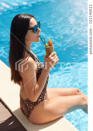 Profile of cheerful young female with darlk long hair in swimming suit with leopard print, drinking cocktail, enjoying resting at pool. Woman sitting on edge of pool with beverage. Vacation concept. 101140011