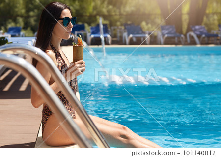 Stylish lady resting at resort, sitting near swimming pool with legs in water on vacation, attractive woman dressed fashionable swimsuit and black sunglasses, looking at distance. Recreation concept. 101140012