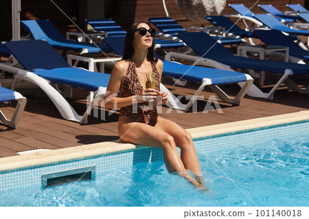 Close up of slim tanned woman sitting on swimming pool edge with cocktail in hand, dressed swimming suit with leopard print and dark sunglasses, lady looking up sky. Recreation and luxury concept. 101140018