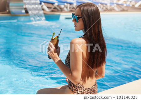 Back view of young pretty woman drinking cocktail against outdoor pool. Female with perfect tanned skin sitting near swimming pool, visiting sea resort. Concept photoof recreation and tourism. Back view of young pretty woman drinking cocktail against outdoor pool. Female with perfect tanned skin sitting near swimming pool, visiting sea resort. Concept photoof recreation and tourism. 101140023