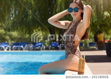 Horizontal outdoor shot of relaxed attractive female wearing leopard swimming suit and sunglasses, touching her hair, sitting near swimming pool, spending time outdoors. Summer and vacation concept. 101140026