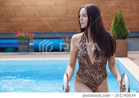 Outdoor shot of thoughtful pensive woman looking aside after swimming in swimming pool, wearing leopard swimming suit, holding onto handrails, spending free time at resort complex, being dreamy. 101140034