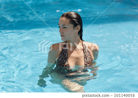 Beautiful woman relaxing in swimming pool in blue clear water. Girl with healthy tanned skin posing in swimming pool, looking aside, being with wet hair, enjoying summer days at luxury resort. Beautiful woman relaxing in swimming pool in blue clear water. Girl with healthy tanned skin posing in swimming pool, looking aside, being with wet hair, enjoying summer days at luxury resort. 101140058
