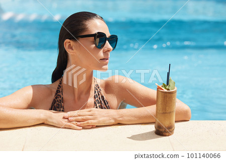 Close up portrait of young woman in swimsuit with leopard print relaxing with fresh drink standing near poolside outdoors, looks aside, has thoughtful facial expression. Summer time and resort concept Close up portrait of young woman in swimsuit with leopard print relaxing with fresh drink standing near poolside outdoors, looks aside, has thoughtful facial expression. Summer time and resort concept 101140066