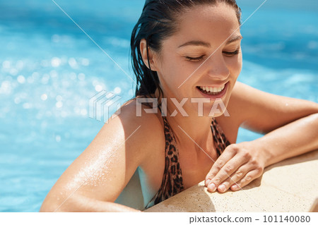 Close up portrait of cheerful good looking brunette swimming in clean water, having rest at swimming pool, smiling sincerely, closing her eyes with pleasure, wearing leopard swimming suit. Rest. 101140080