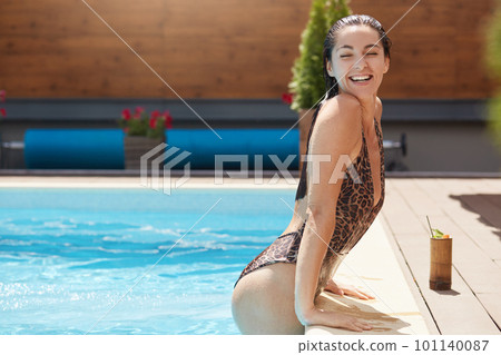 Side view of young woman relaxing in swimming pool. Beautiful Caucasian female with wet hair in swimsuit with leopard print enjoys sunny days at resort. Outdoor hotel pool and summer vacation concept 101140087