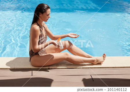 Summer holiday, fashion, vacation concept. Outdoor shot of young beautiful woman with longslim lags on wooden pier at pool applying sunscreen on pefect body, female wearing swimsuit with leopard print 101140112