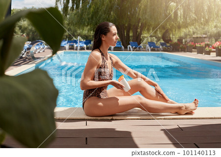 Portrait of attentive focused woman posing near swimming pool over chaise lounge background, being wet after swimming, enjoying vacation time, applying sunbathing cream to skin. Spa resort concept. 101140113