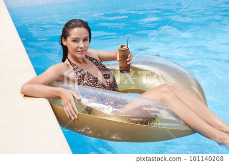 Outdoor shot of young brunette woman with wet hair relaxing on inflatable swim ring, wearing fashion swimwear with leopard print, girl enjoying coctails in swimming pool during summer vacation. 101140208
