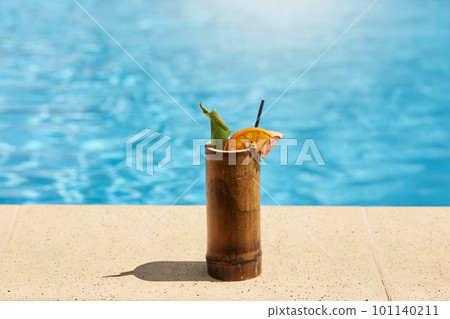 Exotic cocktail in wooden glass with lemon and drinking tube standing on poolside with blue pool's water on background, tasty fresh beverage on resort. 101140211