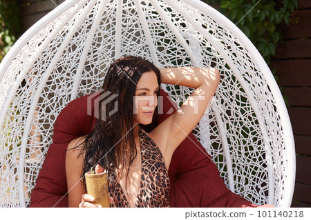 Beautiful attractive young brunette woman in stylish swimming suit with leopard print, posing in white hanging chair, lady enjoying having rest and drinking cocktail. 101140218