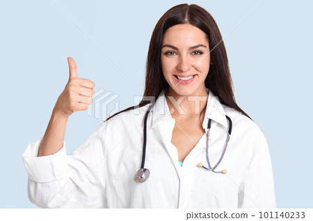 Horizontal shot of pretty talented female doctor with satisfied expression, raises thumb, shows like gesture, approves something, likes her job and helping people, isolated over blue background 101140233
